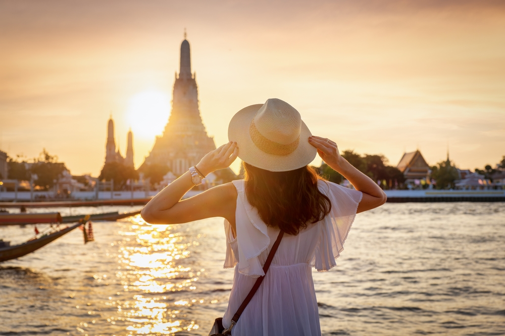 A tourist woman enjoys the view to the famous Wat Arun temple in Bangkok, Thailand, during golden sunset time